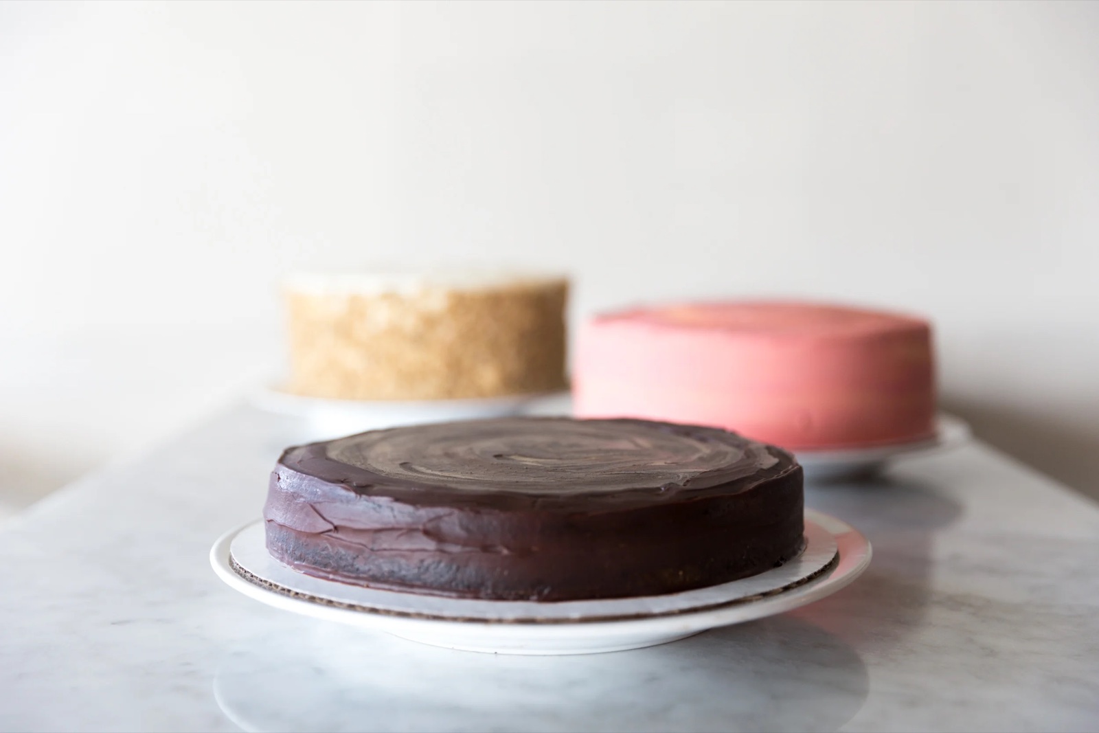 Three frosted cakes on marble: dark chocolate ganache, walnut-crusted and pink.