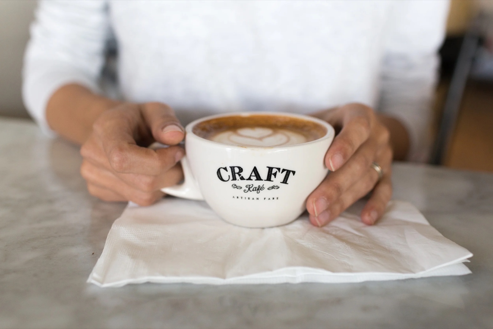 Two hands cradling a Craft Kafe cappuccino with leaf latte art on a marble table.